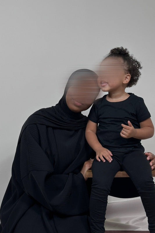 abaya yana noire zoom sur les détails du tissu de la robe pour allaiter et le mannequin pose avec un enfant à ses côtés